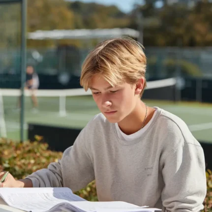 Teenager reading his textbook. There is a tennis court in the background