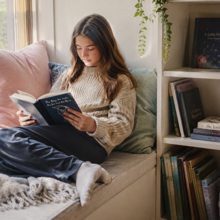 Child reading a book in a cozy window nook at home with bookshelves nearby.
