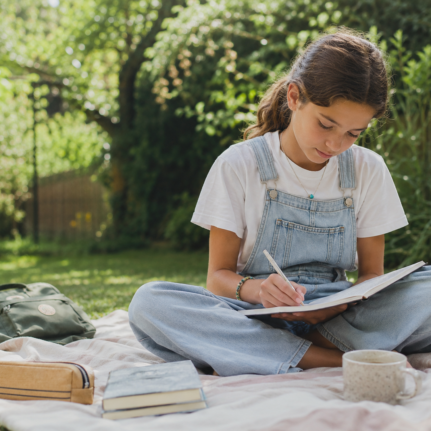 Child sitting on a picnic blanket in the garden, writing in a journal with books and a backpack nearby.